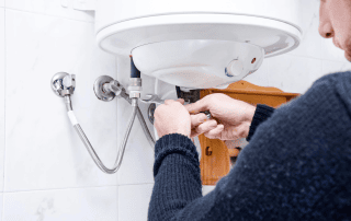 A person uses a wrench to repair or install pipes under a wall-mounted white water heater in a bathroom with white tiled walls.