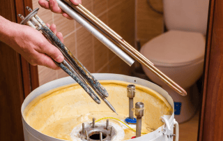 A person holds new and corroded heating elements above an open water heater tank, showing the difference in condition. A toilet and bathroom tiles are visible in the background.